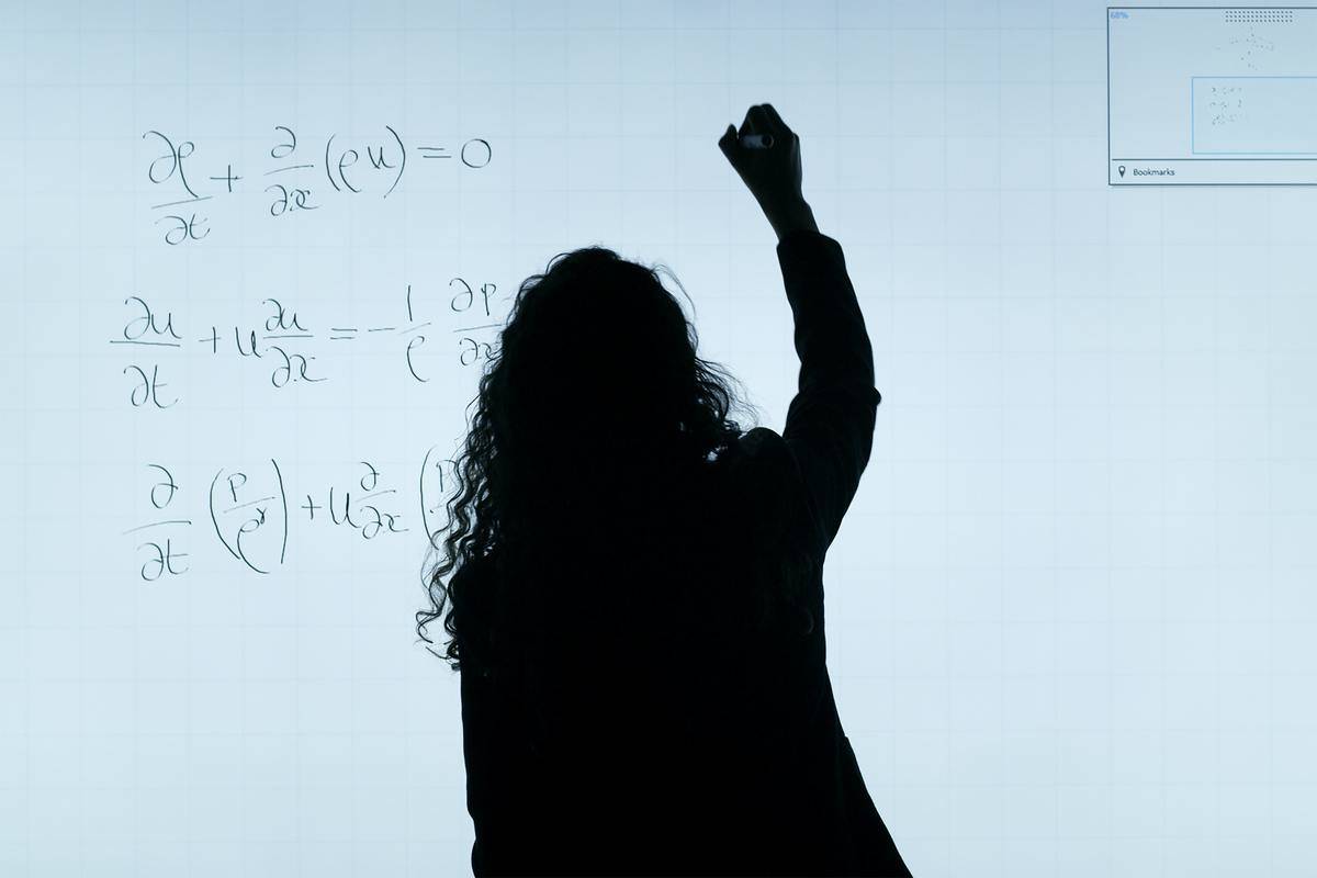 A silhouette of a woman writing out equations on a white board.