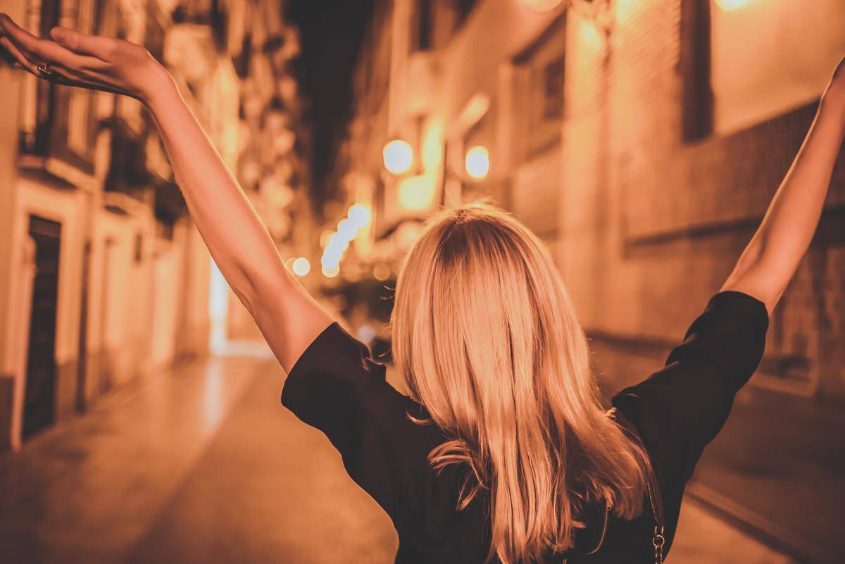 A woman walking down a lit street with her arms up in a carefree fashion.