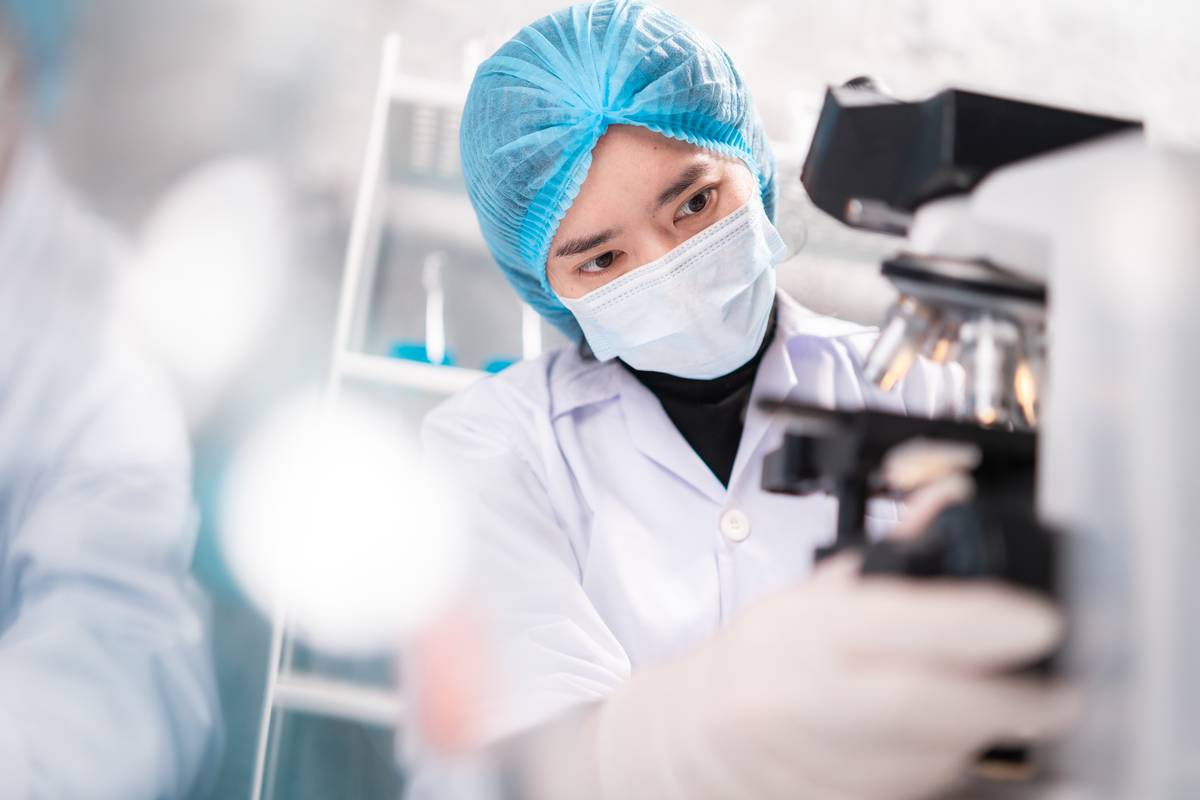 A researcher in PPE examining findings under a microscope.
