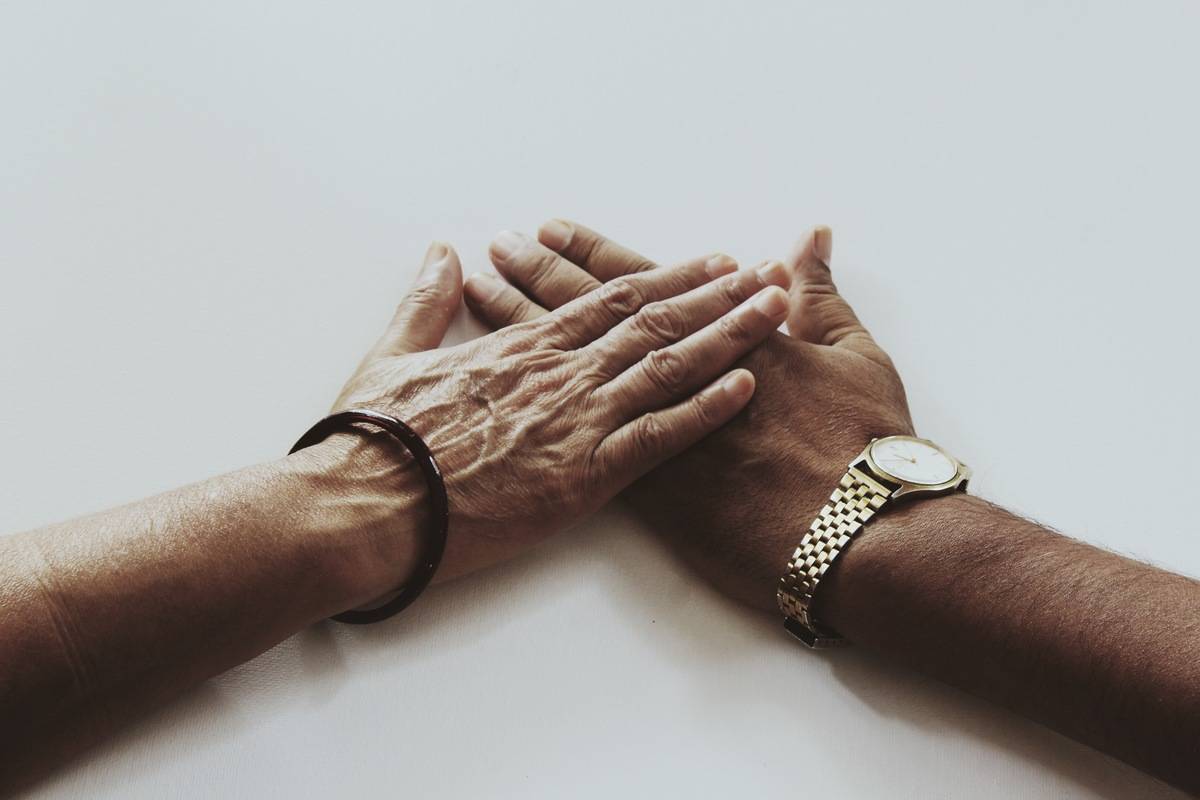 Two hands, one more elderly than the other, placed on top of one another on a white tabletop.