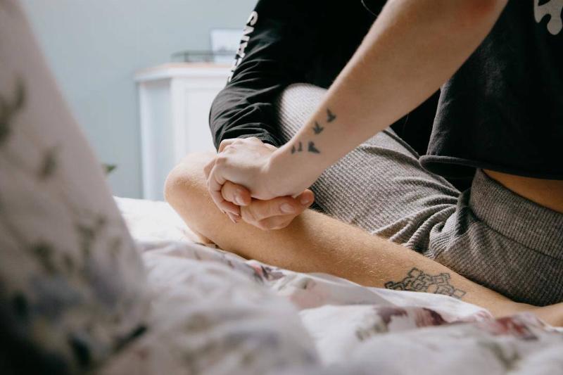 tatooed couple sitting cross-legged together on a bed, holding hands.