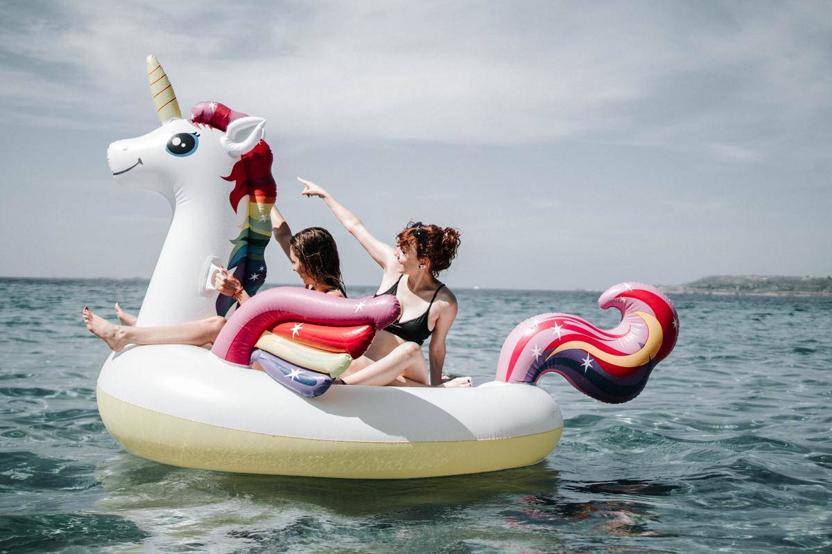Two women smiling as they ride on a large, rainbow unicorn pool floatie.