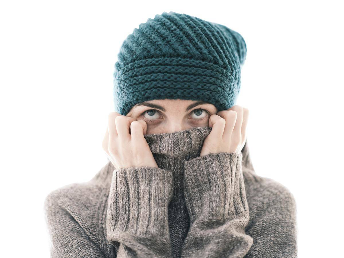 A woman in a sweater and hat, looking tired, pulling the sweater cowel over her mouth.