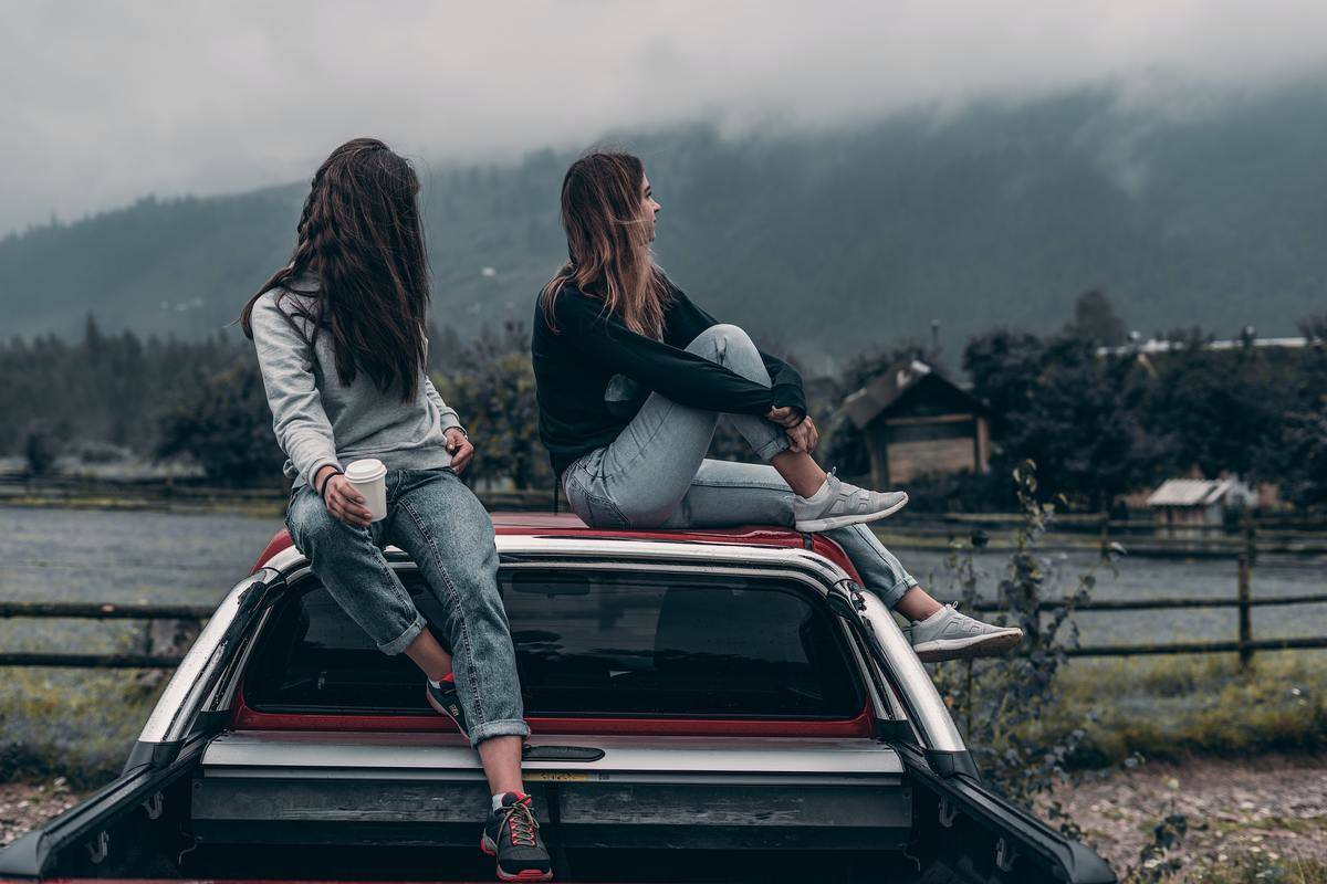 Two girl sitting on top of a car, looking off into the distance.