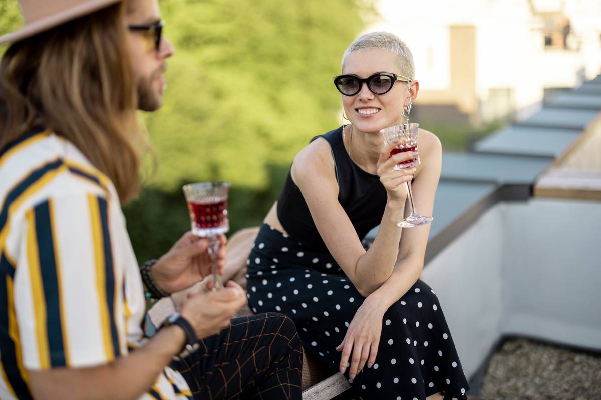 Two stylish carefree persons talk and drink wine at roof top terrace at countryside house. Friends hanging out at picnic outdoors.