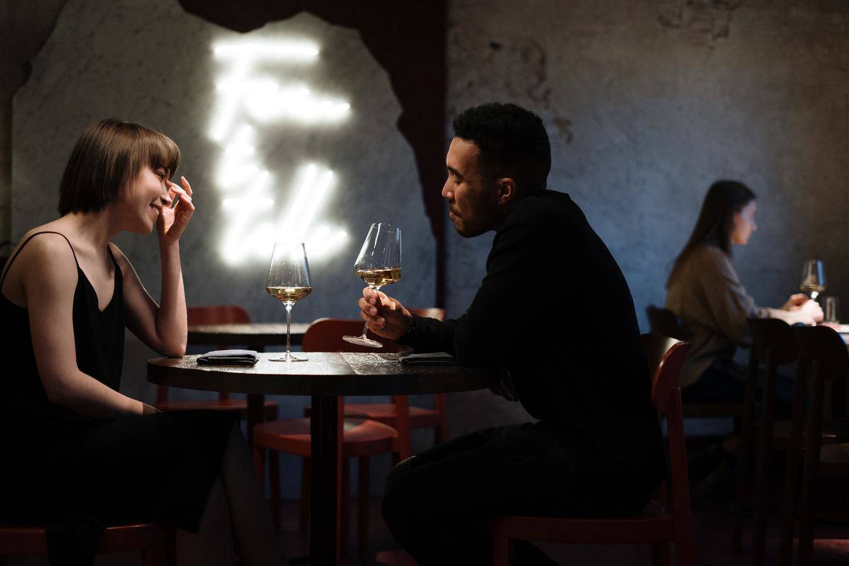 A man and a woman at a table in a restaurant, talking over two glasses of wine.