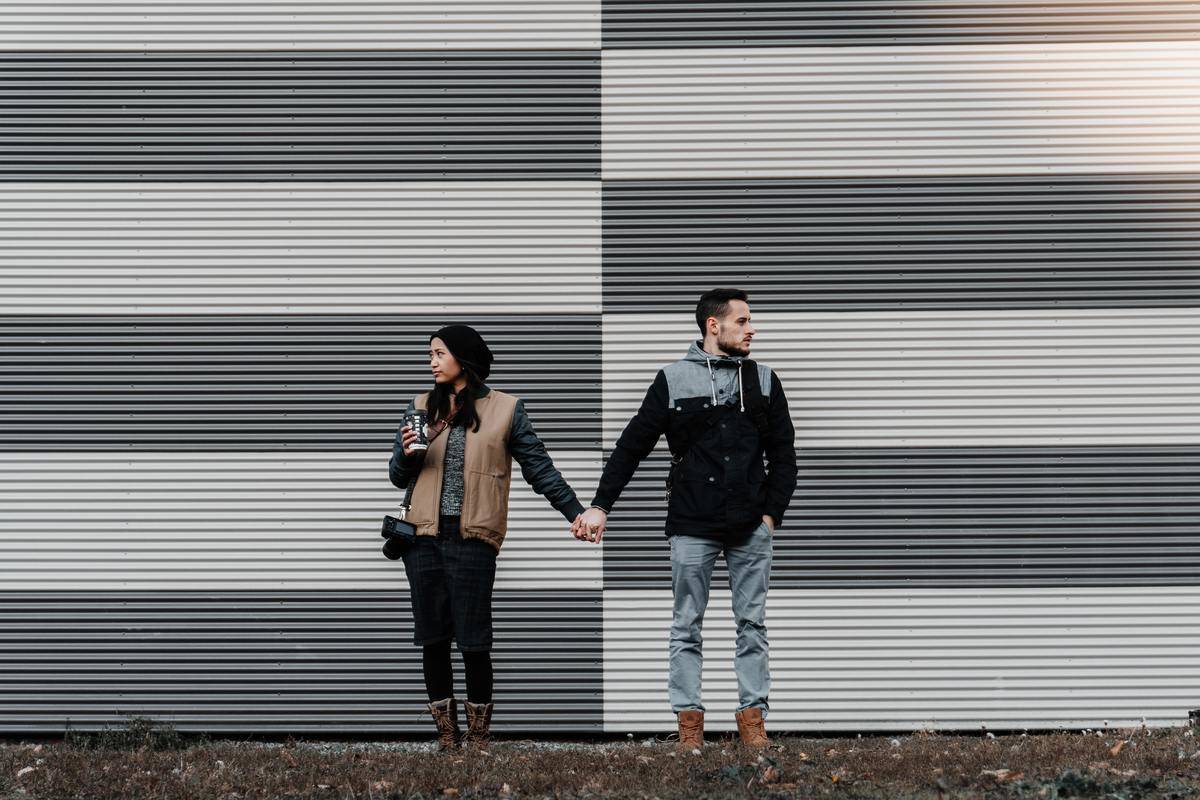 A couple standing in front of a grey and white striped wall, holding hands but looking away from one another.