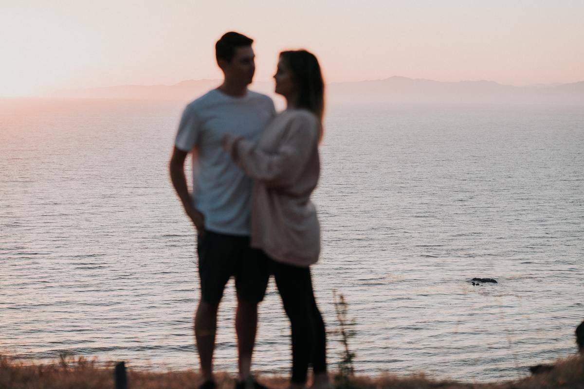 A couple hugging, slightly blurred in the foreground, the water behind them.