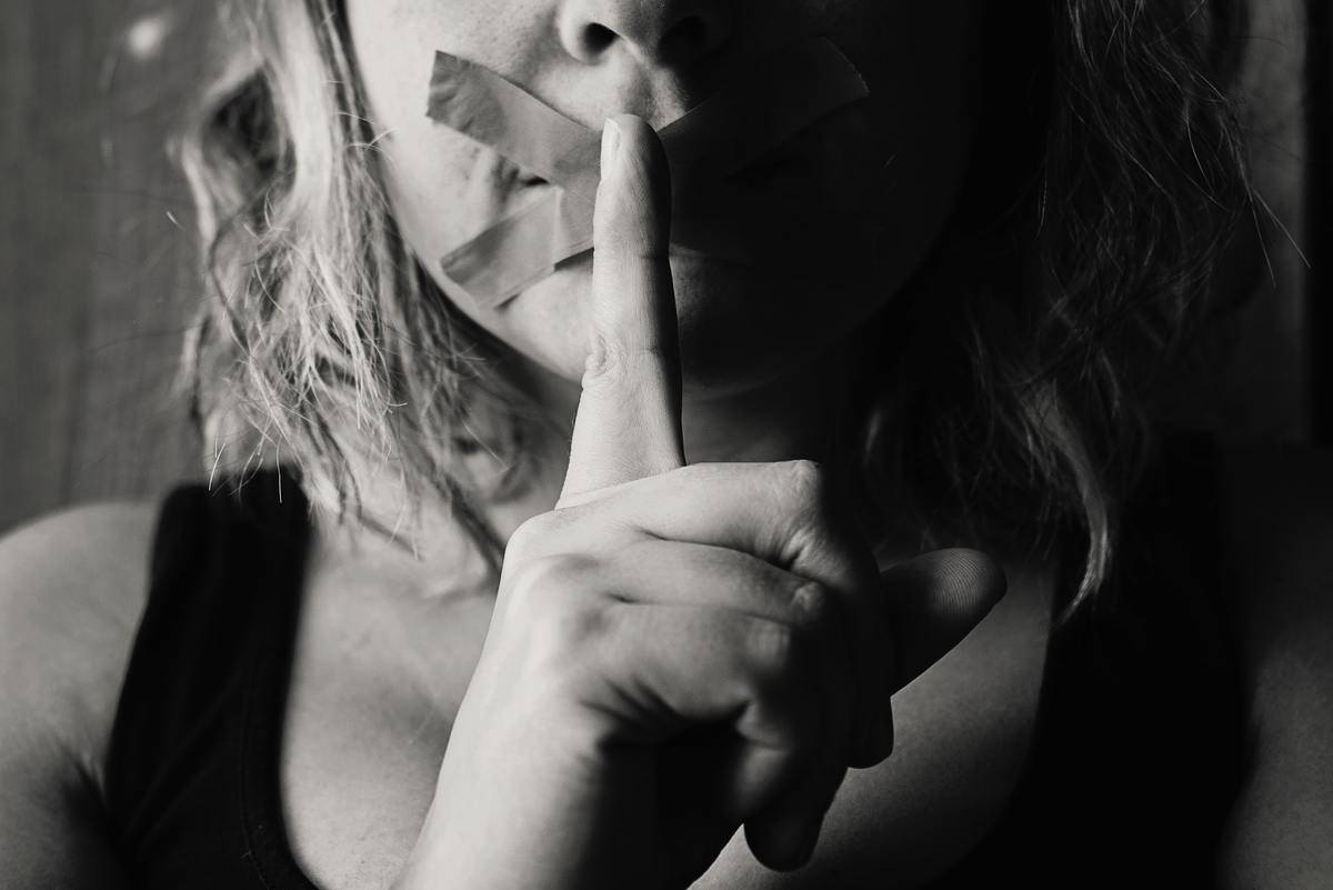 A woman with tape over her mouth in an 'x' shape also holding a finger in front of her lips.