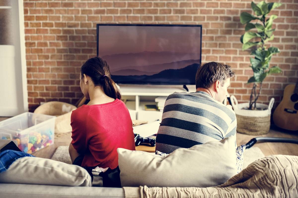 A couple sitting on a couch in their living room, turned away from one another.