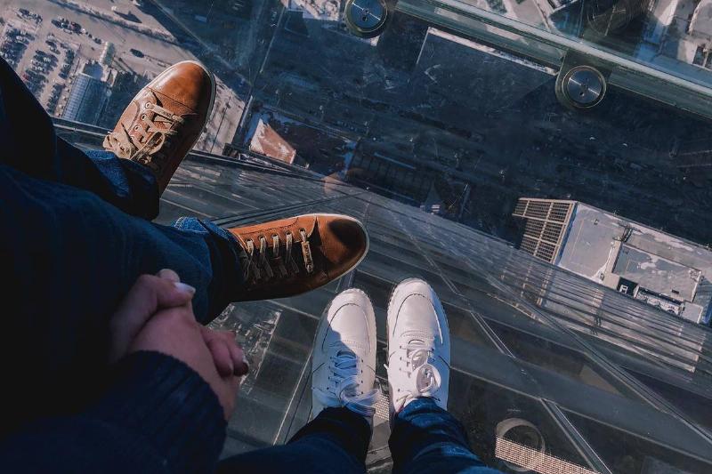 A couple holding hands, looking down through a glass floor at city streets below.