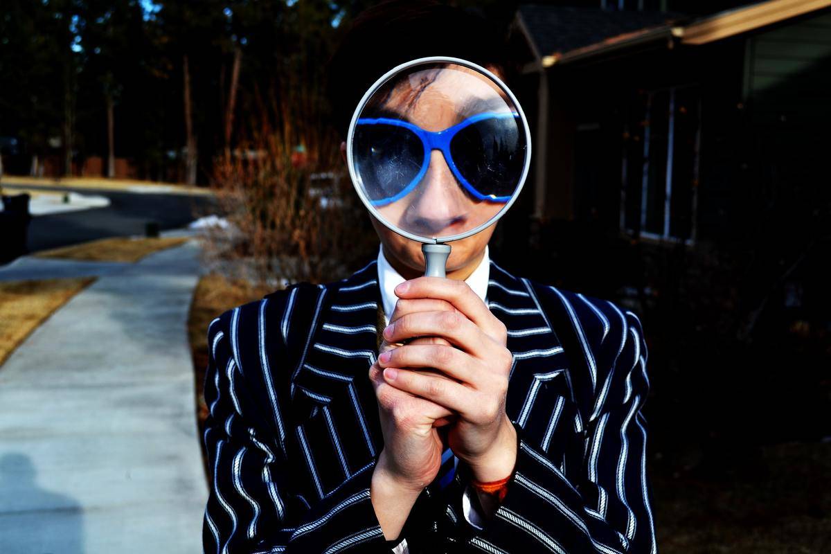 A person in blue sunglasses and a striped suit holding up a magnifying glass in front of their face.