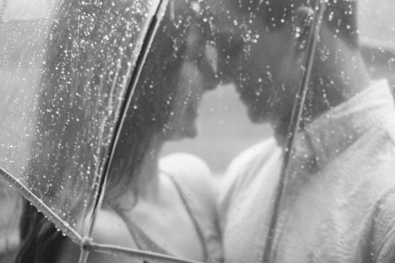 A greyscale image of a couple embracing, seen through a transparent umbrella.