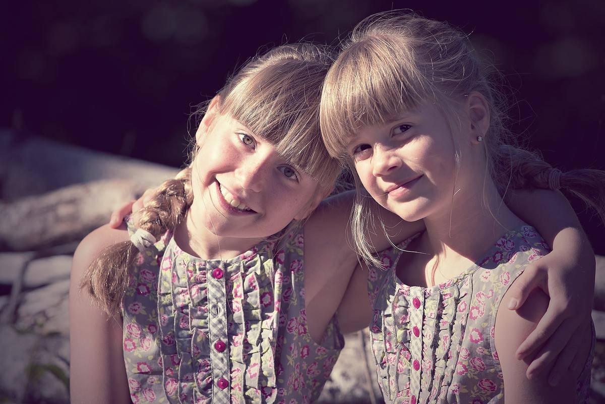A set of young female twins, wearing matching dresses and smiling at the camera, each eith an arm around each other.