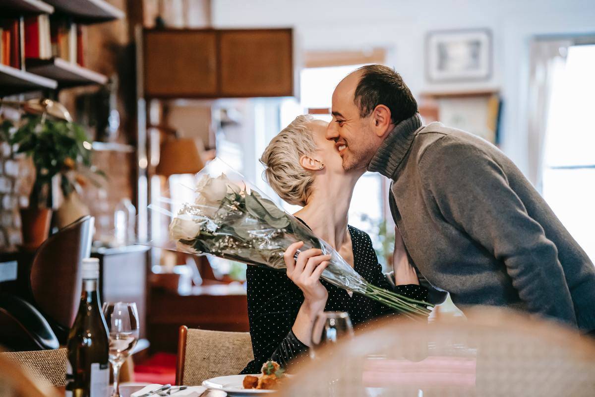 A woman leaning up in her seat to kiss the cheek of a man who just handed her flowers.
