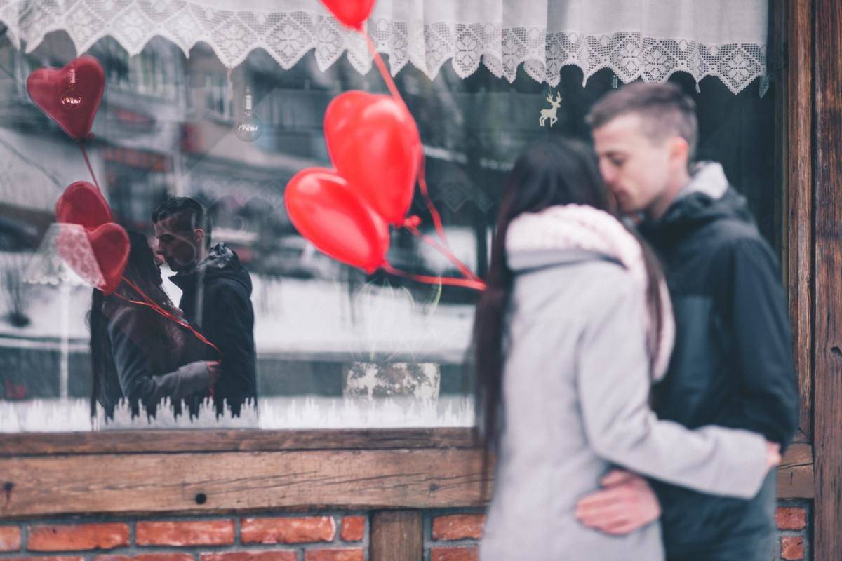 A man and a woman kissing in front of a storefront holing heart balloons.