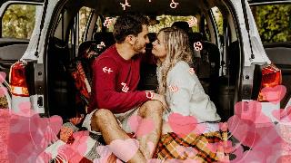 A couple seated in the back of a van, having laid blankets down in the trunk. There are hearts at the bottom of the image as well as a ring of the zodiac symbols around their heads.