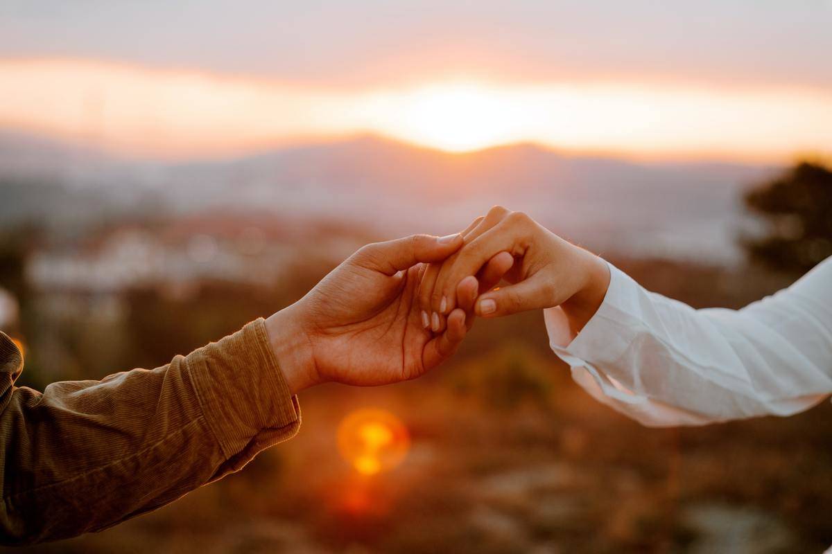 Two people delicately holding hands in front of a sunset.