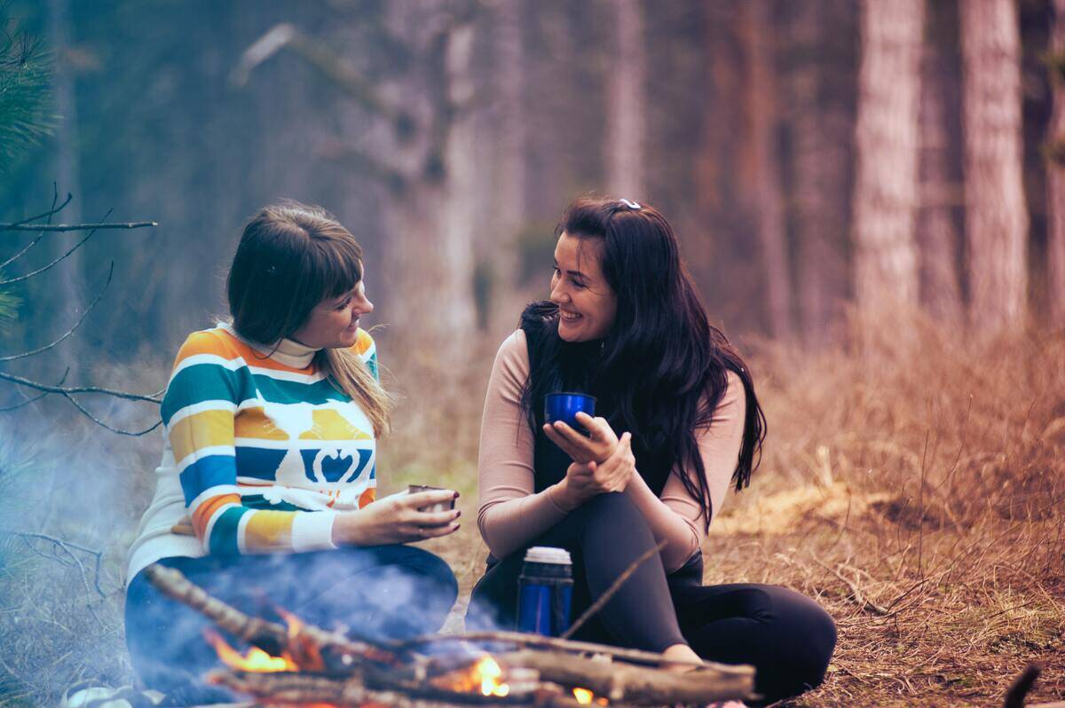 Two friends out camping, smiling as they talk by a fire.