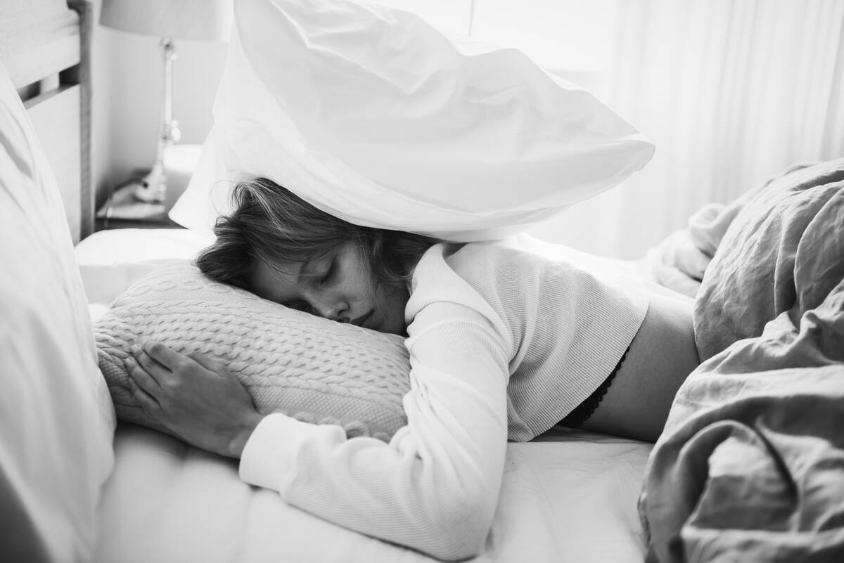 A greyscale image of a woman laying in bed, a pillow on top of her head.