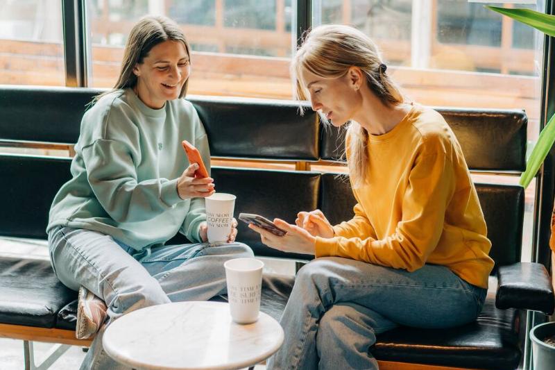 Two cheerful laughing women friends in a coffee shop chatting while showing each other their phones.