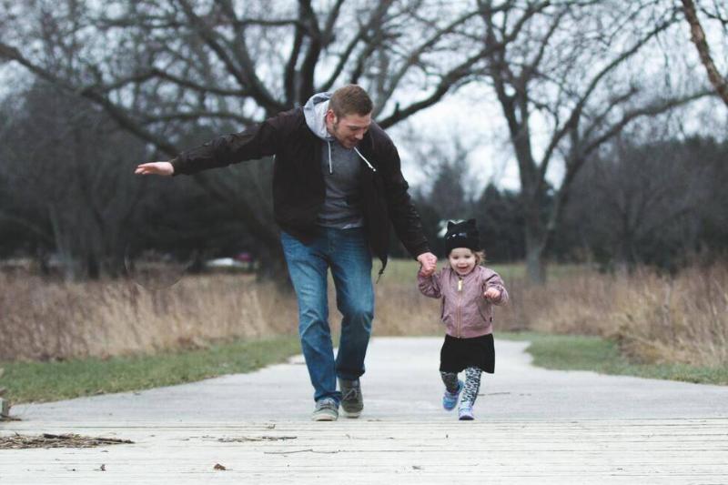 A man holding hands with his young daughter as they run down a path.