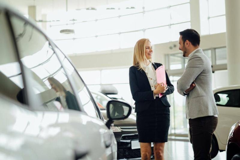 A female car salesperson speaking to a male client on a show floor.