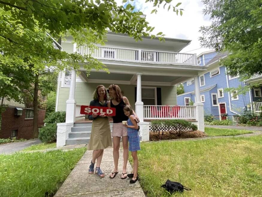 Two of the women and one kid standing in front of the house with the 'sold' sign.