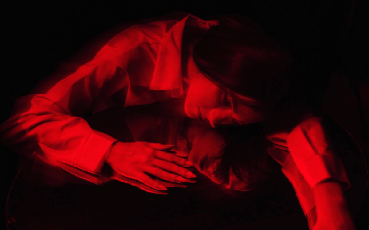 A woman with sharp nails laying on top of a mirror, lit in red in the dark, looking at her reflection.