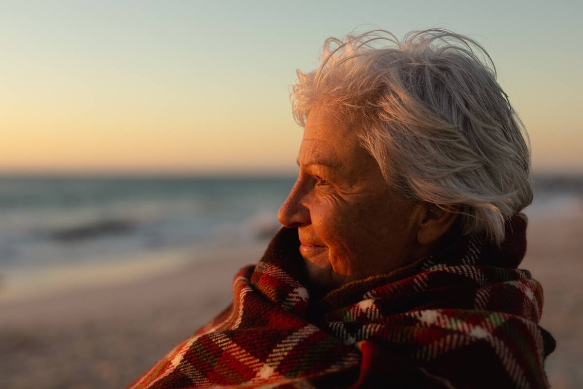 Side view close up of a senior woman standing on the beach at sunset smiling and looking out to sea.