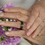 Elderly couple holding hands and showing their wedding rings and bouquet, constellations decorating the corners of the image.