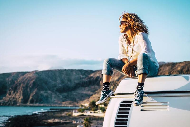 Woman sitting down on the roof of a old nice vintage camper van in front of some cliff ranges and water.