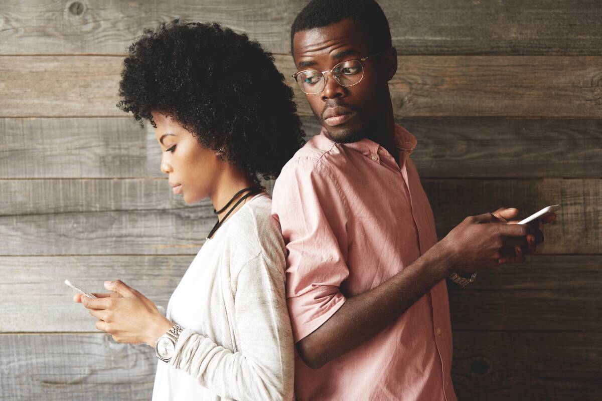 Couple standing back-to-back, holding mobile phones. Man looking over his shoulder at his girlfriend trying to see what she is texting and who she is messaging.