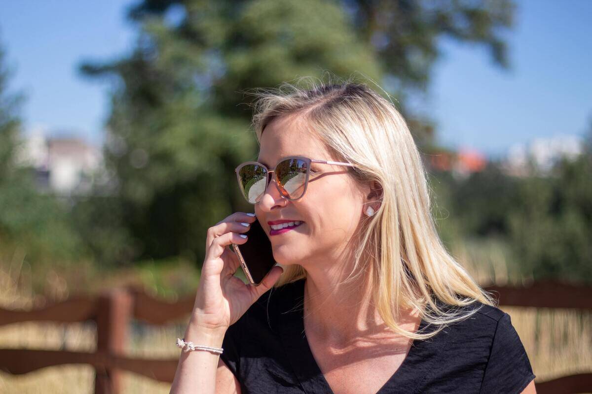 A woman in sunglasses, standing outside, talking on her cell phone.