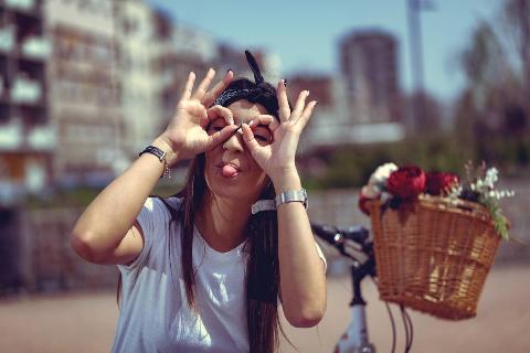 Pretty happy young woman is enjoying and having fun showing tongue, in a summer sunny day, beside the bike with flower basket.