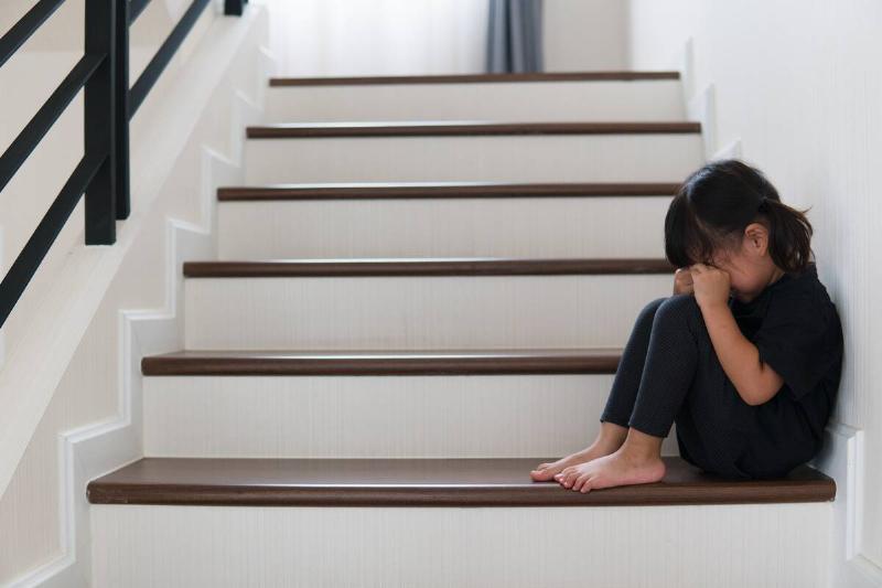 A young child sitting on a stair, knees to her chest, crying.