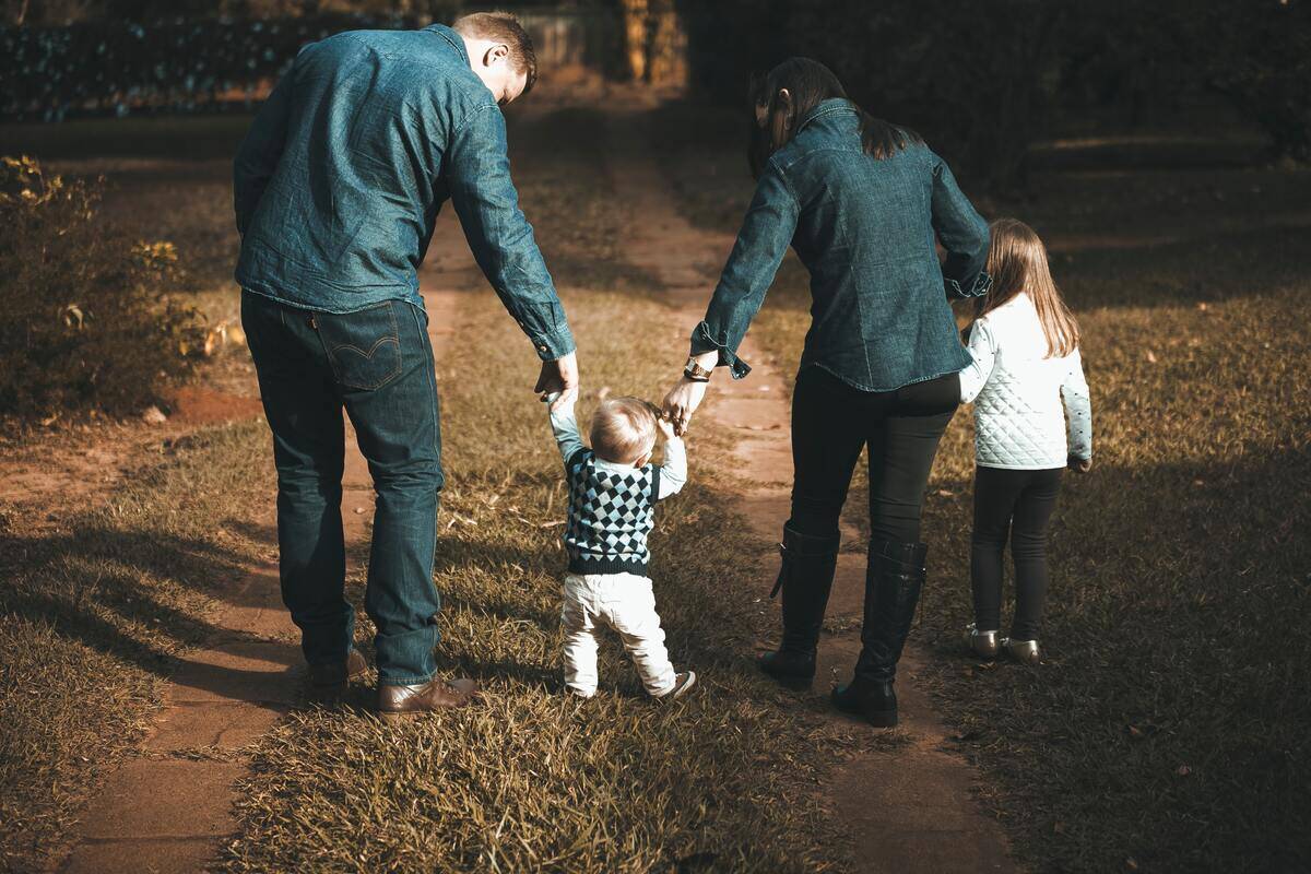 Two parents each holding one of their baby's hands, helping him walk.