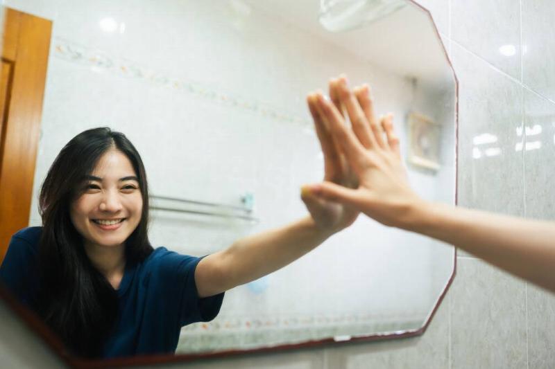 A woman smiling at herself in a mirror, a hand reaching out to touch its reflection.