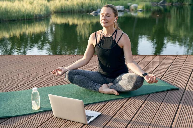 Sporty relaxed woman meditating, sitting in Lotus pose while watching tutorial on a laptop, doing yoga on a mat in nature on a sunny day outdoors.