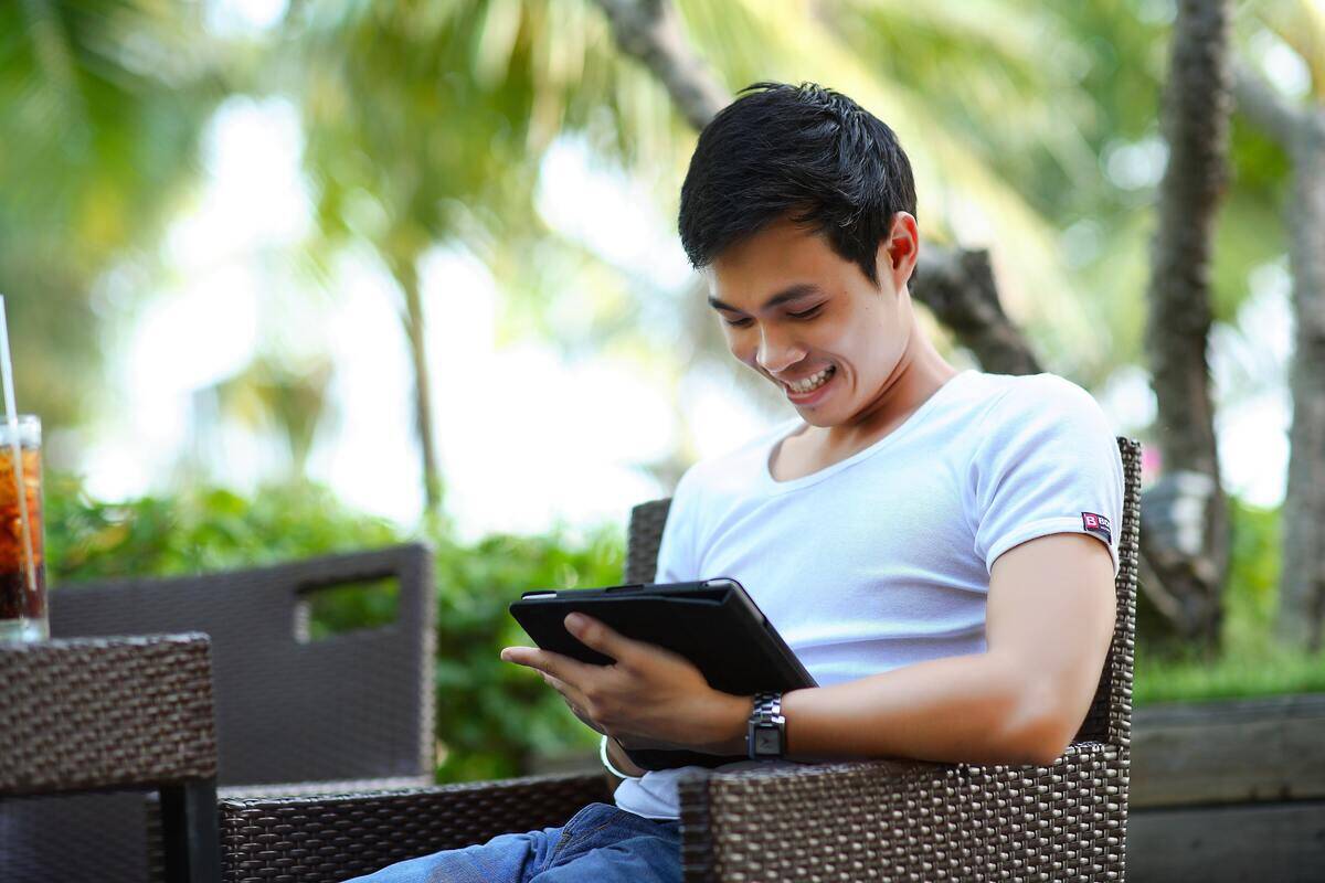 A man sitting outside, smiling as he looks down at a tablet.