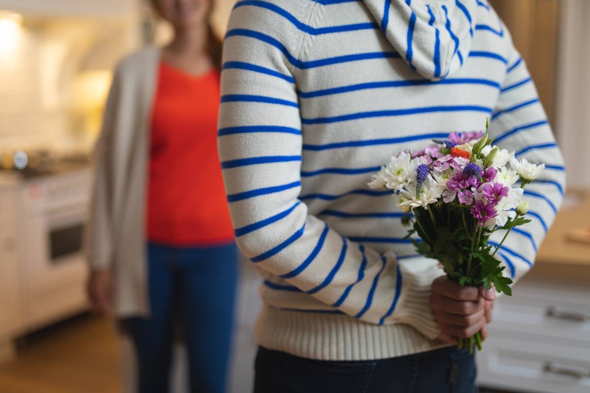 Rear view mid section of a Caucasian man at home, wearing a blue striped sweater and holding a bunch of flowers behind his back, his female partner standing in the background facing him, focus on the foreground