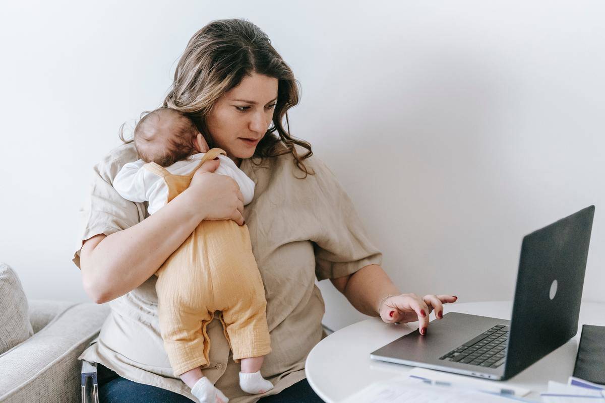 A woman holding a baby against her chest in one arm, using her laptop with the other hand.