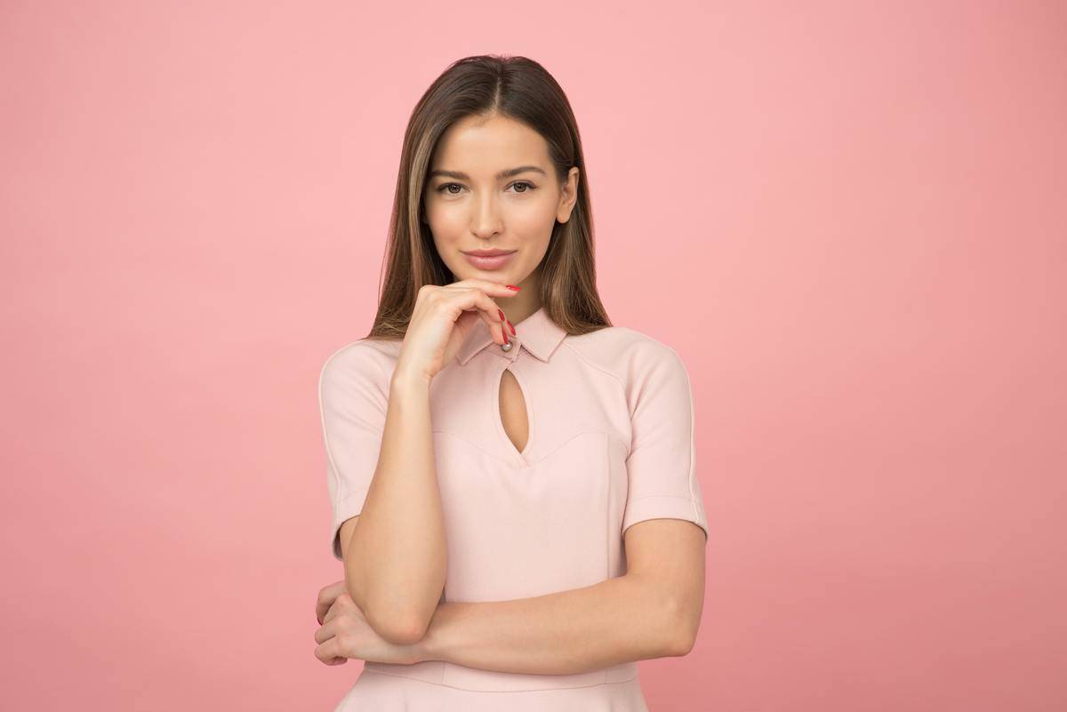 A confident woman looking at the camera, hand poised under her chin, wearing a pink dress against a pink background.