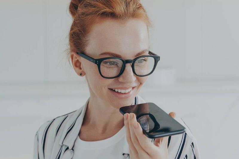Close up shot of redhead woman in glasses speaking into her phone's microphone while smiling.
