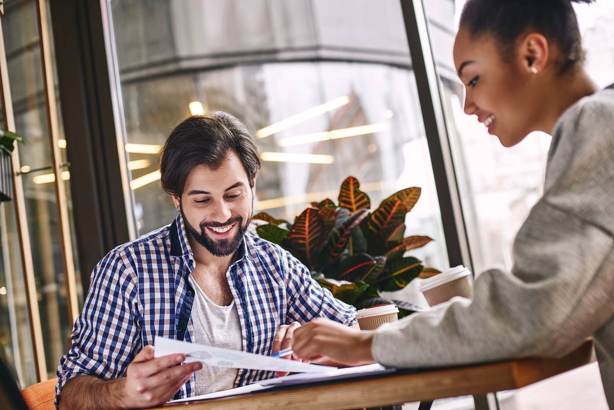 Two cheerful colleagues discussing new ideas sitting over desk. Bearded caucasian man and attractive african american woman look on charts.