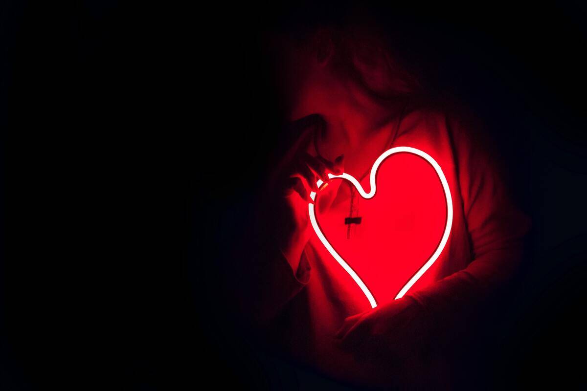 Someone holding a red neon light in the shape of a heart to their chest.