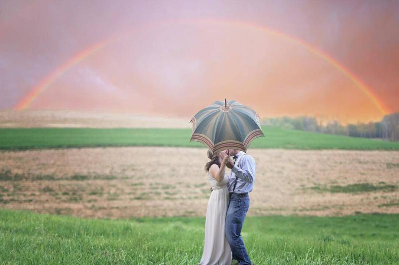 Two people kissing behind an umbrella in a field, standing below a rainbow.