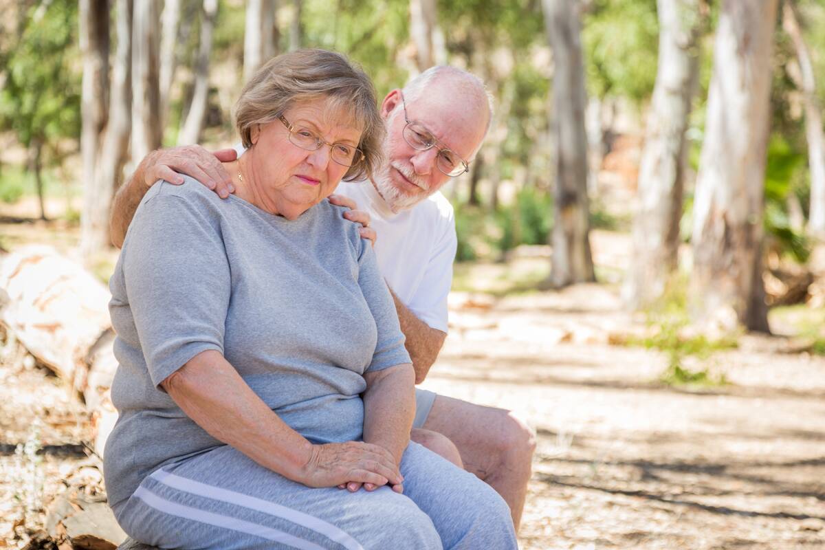 Very Upset Senior Woman Sits With Concerned Husband Outdoors, her husband's hands on her shoulders.