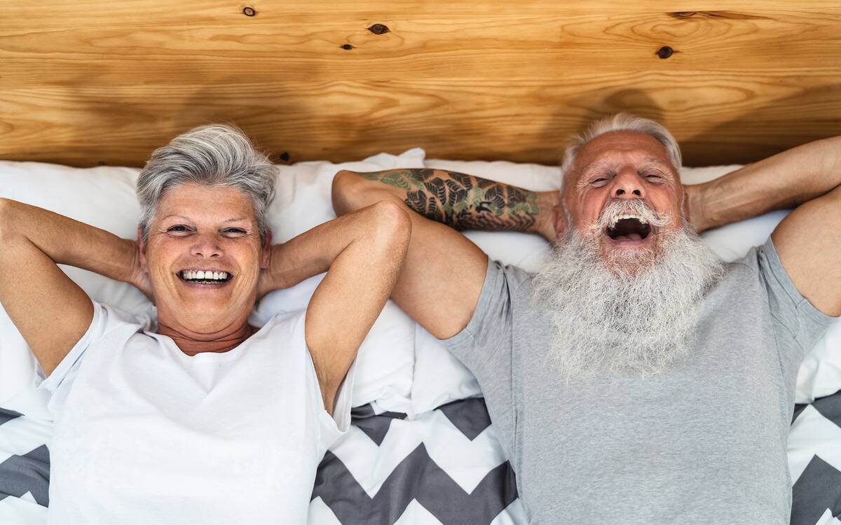 A couple smiling as they lay in bed side-by-side, hands behind their head. 