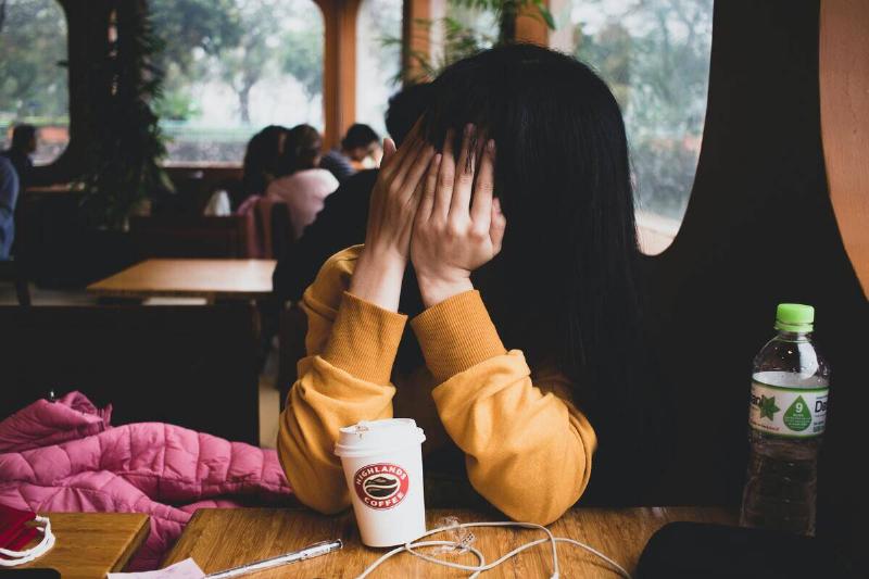 A woman seated at a cafe, hiding her face from the camera with her hands.